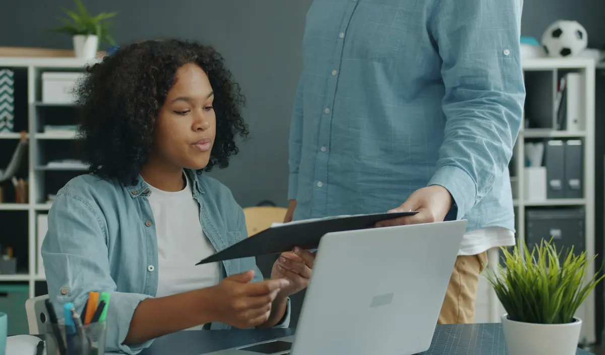 Doi colegi la birou verifică un document, lângă un laptop.